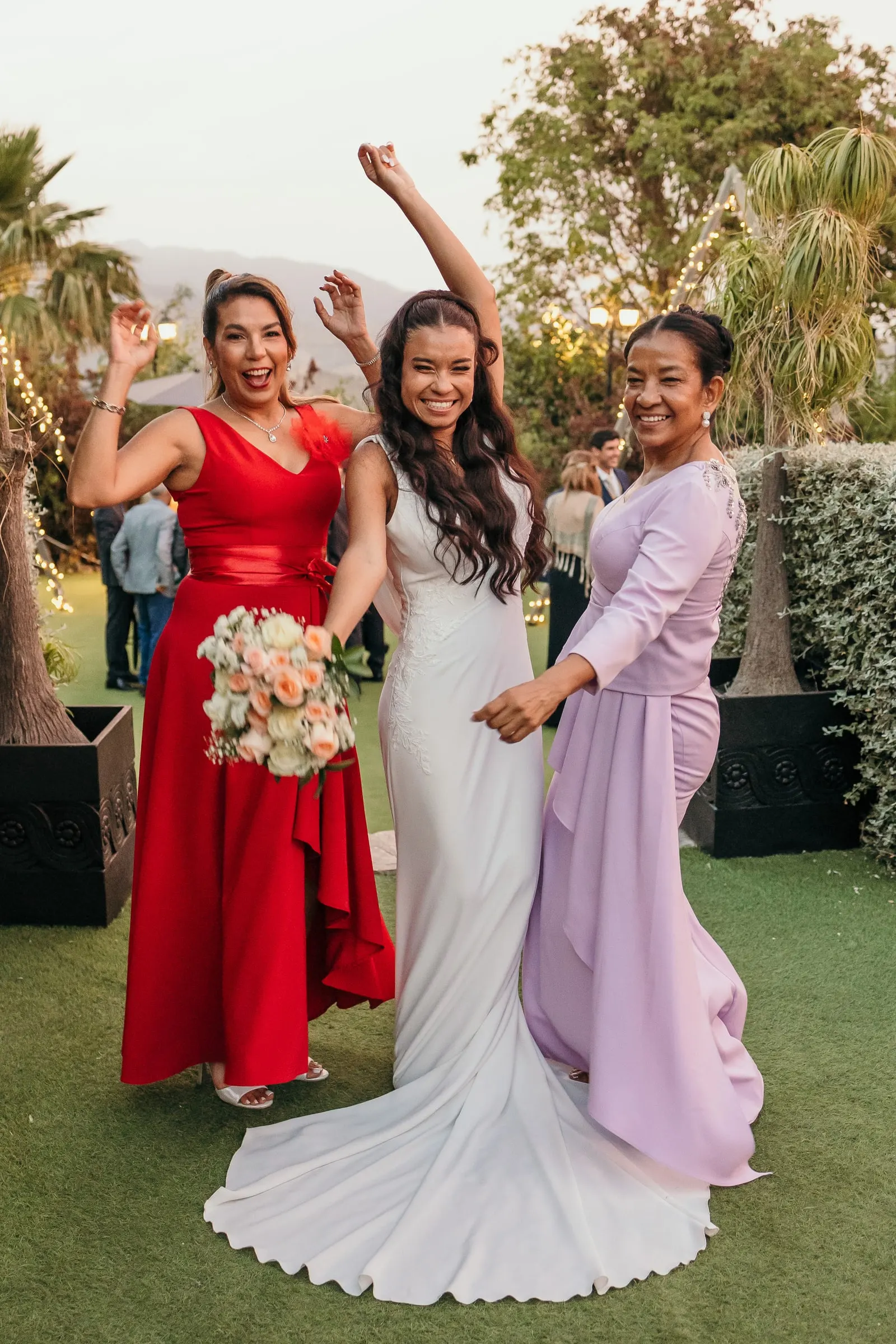 Bride dancing on the lawn with two guests in red and lilac, arms in the air