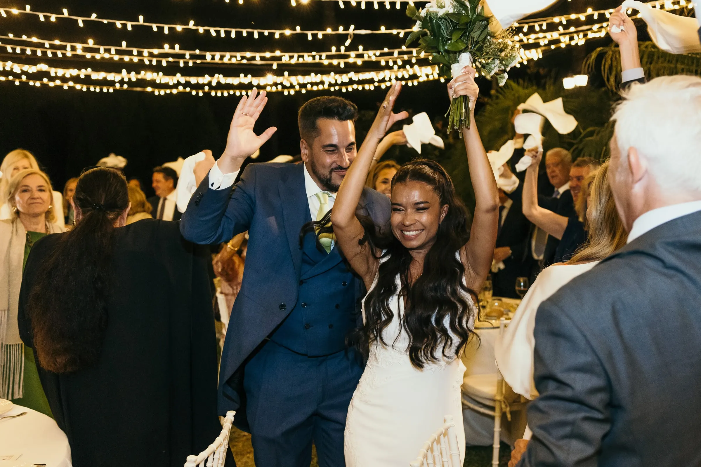 Couple waving white napkins with the guests under strings of fairy lights at the wedding dinner
