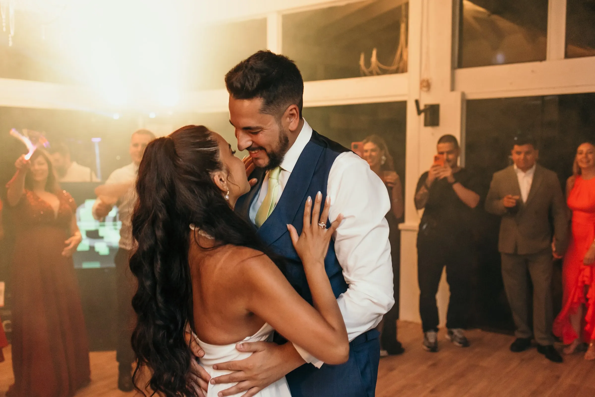 Couple kissing on the dance floor against warm backlight as guests watch